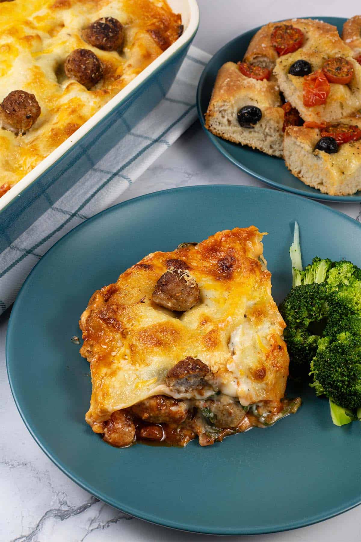 A portion of three cheese meatball lasagne on a teal dinner plate with broccoli on the side and tomato and olive focaccia in the background.