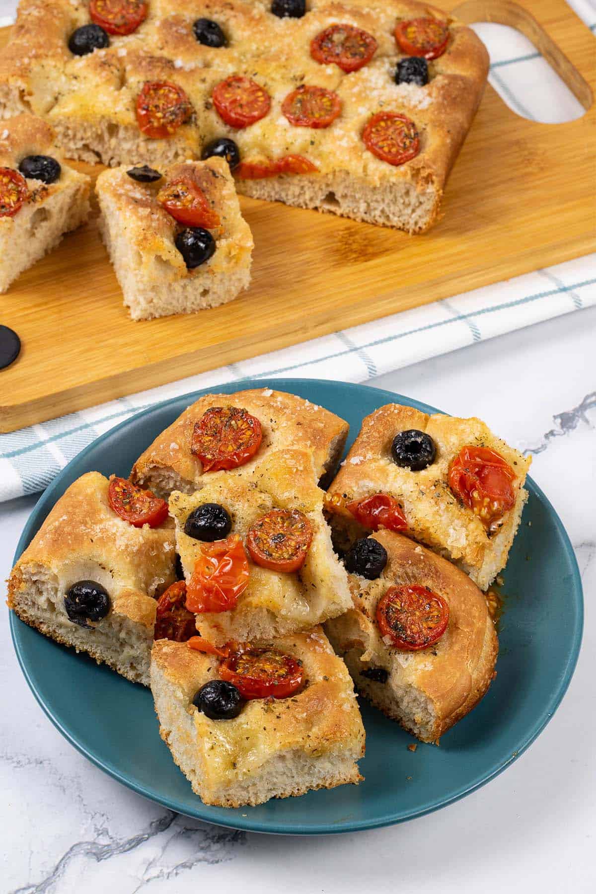 Six slices of tomato and olive focaccia on a teal side plate with the rest of the bread on a board in the background.