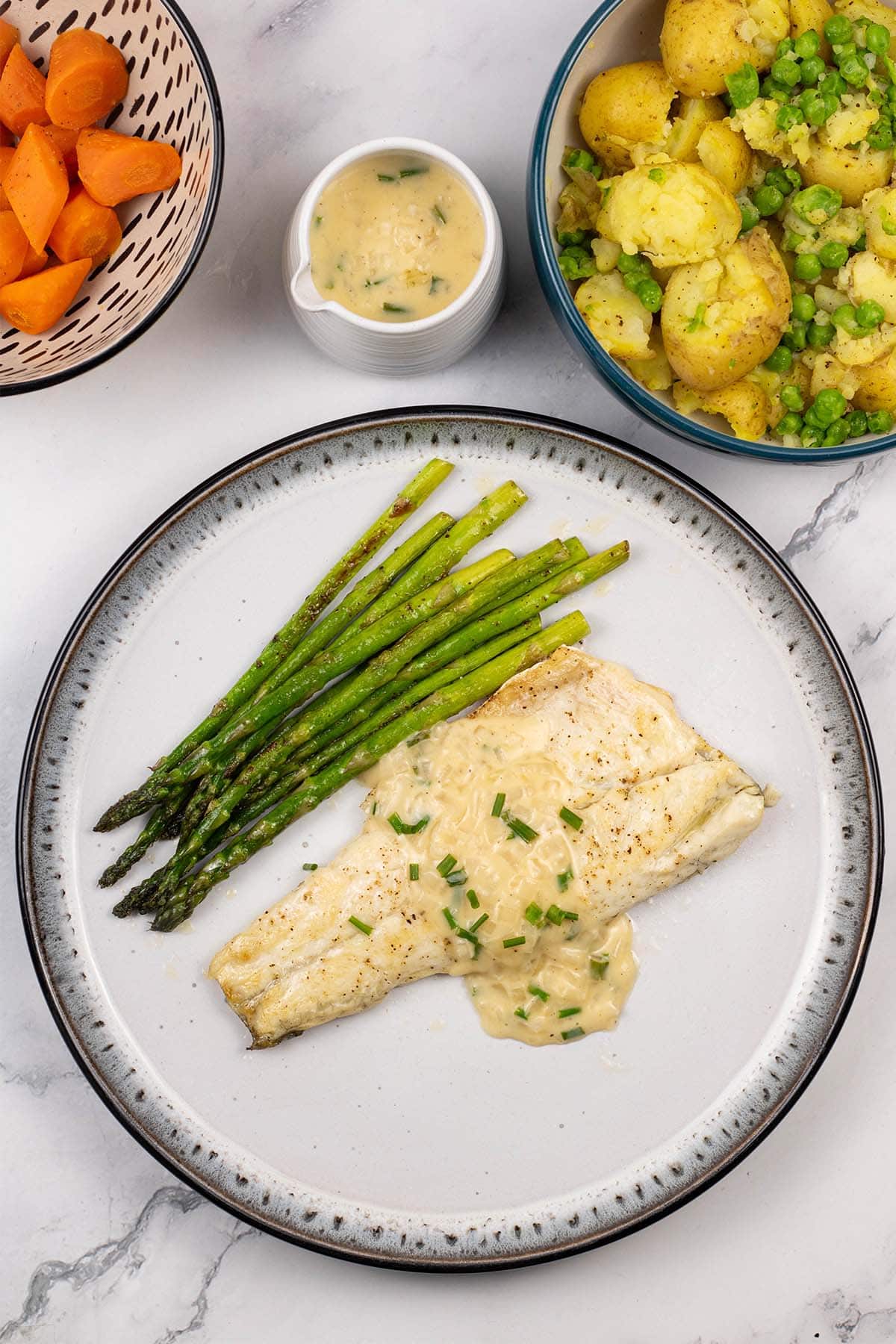 White wine and cream sauce spooned over a sea bass fillet with asparagus on a black and light grey patterned plate.