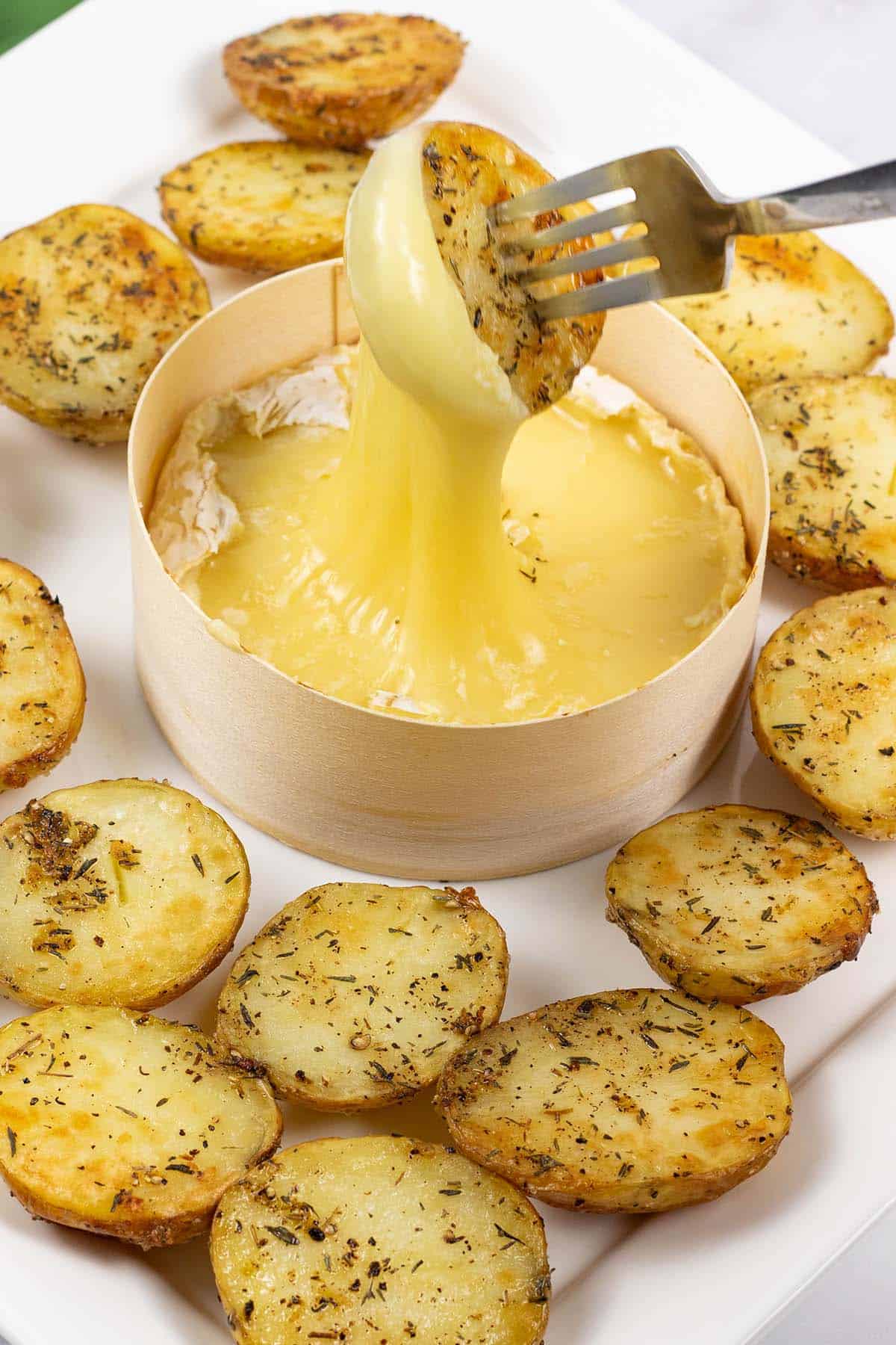 Close up of baked camembert with thyme baby potatoes on a square white platter, with a potato dipped into be oozy cheese!