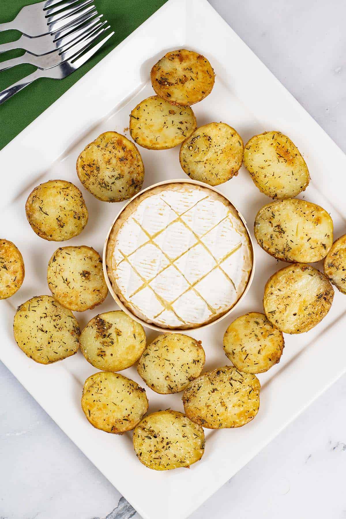 Baked camembert with thyme baby potatoes on a square white platter, with a green napkin and 4 forks on the side.