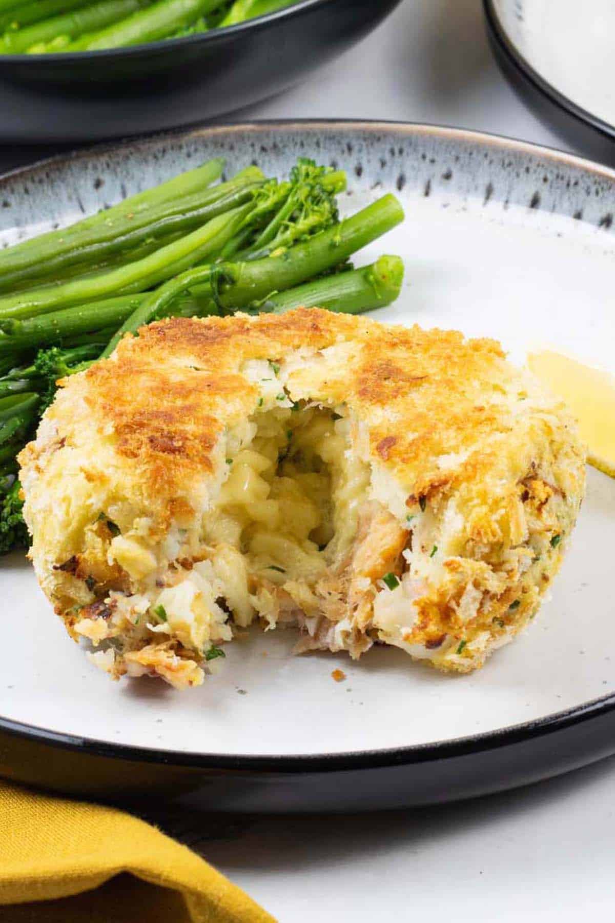 A melt in the middle fishcake on a black and white patterned dinner plate with green beans and tenderstem broccoli, and a wedge of lemon.