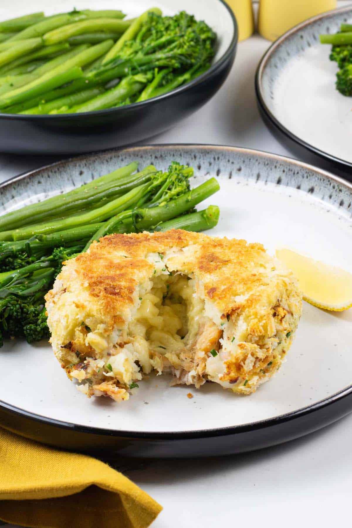 A melt in the middle fishcake on a black and white patterned dinner plate with green beans and tenderstem broccoli, and a wedge of lemon.