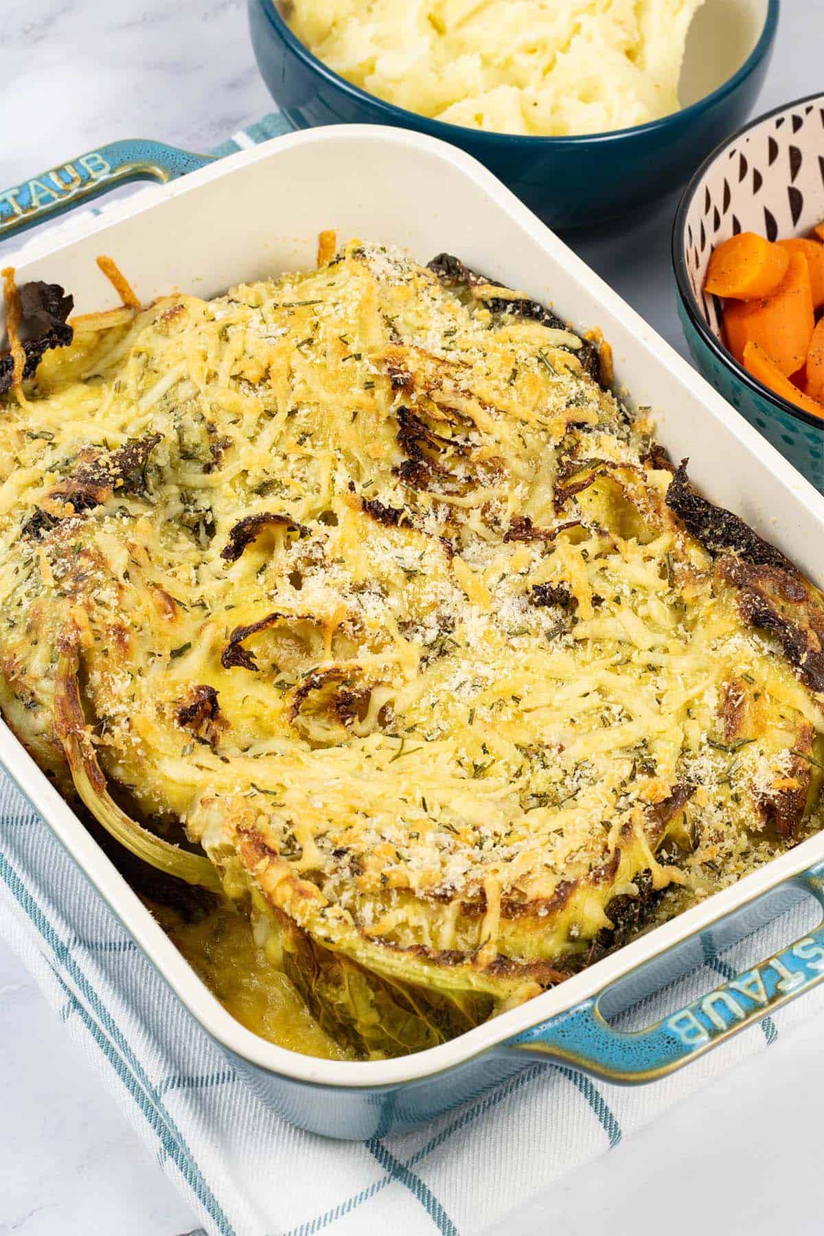 Savoy cabbage gratin with herby cheddar crumbs in a rectangle Staub dish with bowls of mashed potatoes and boiled carrots on the side.