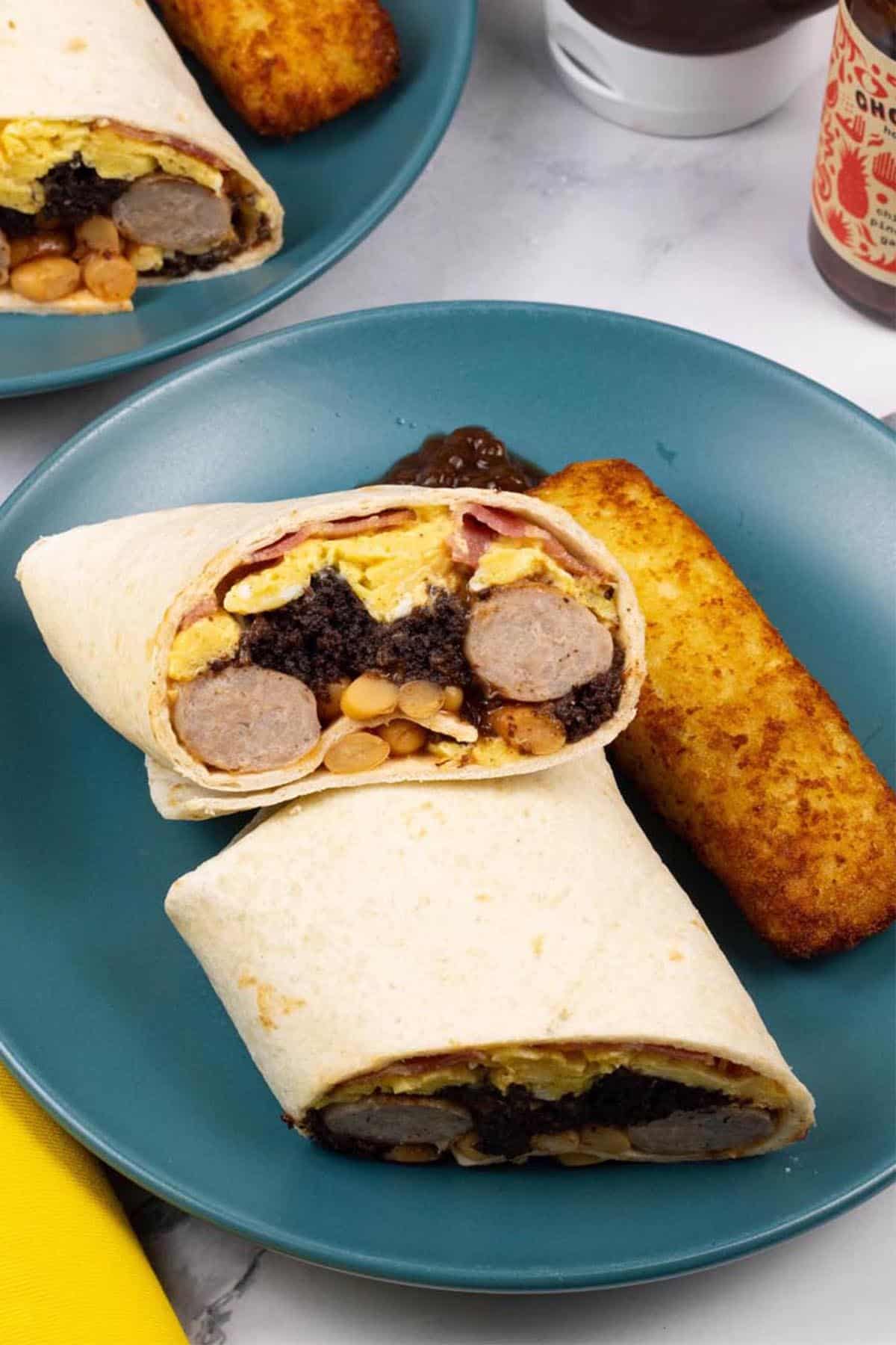 Two portions of breakfast burritos on teal side plates with a cheesy hash brown and blob of brown sauce on the side.