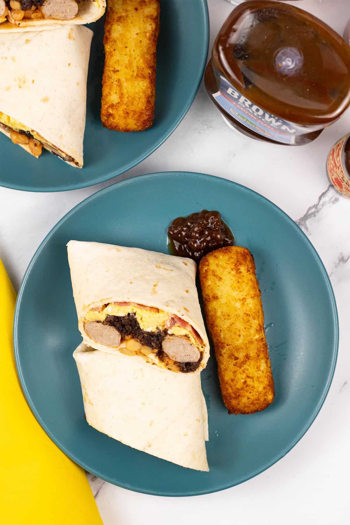 Two portions of breakfast burritos on teal side plates with a cheesy hash brown and blob of brown sauce on the side.