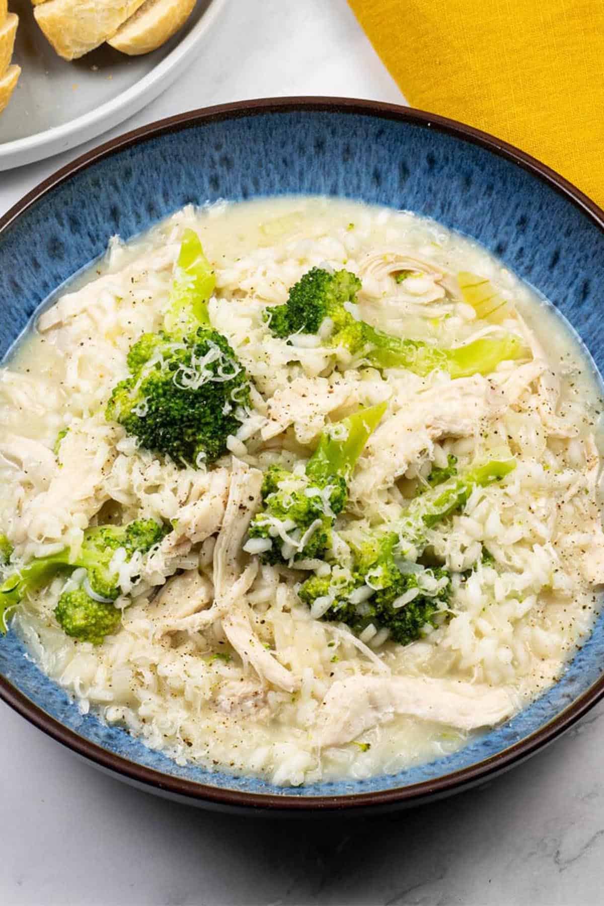 Chicken and broccoli risotto in a blue patterned bowl with a plate of sliced baguette and a mustard napkin on the side.