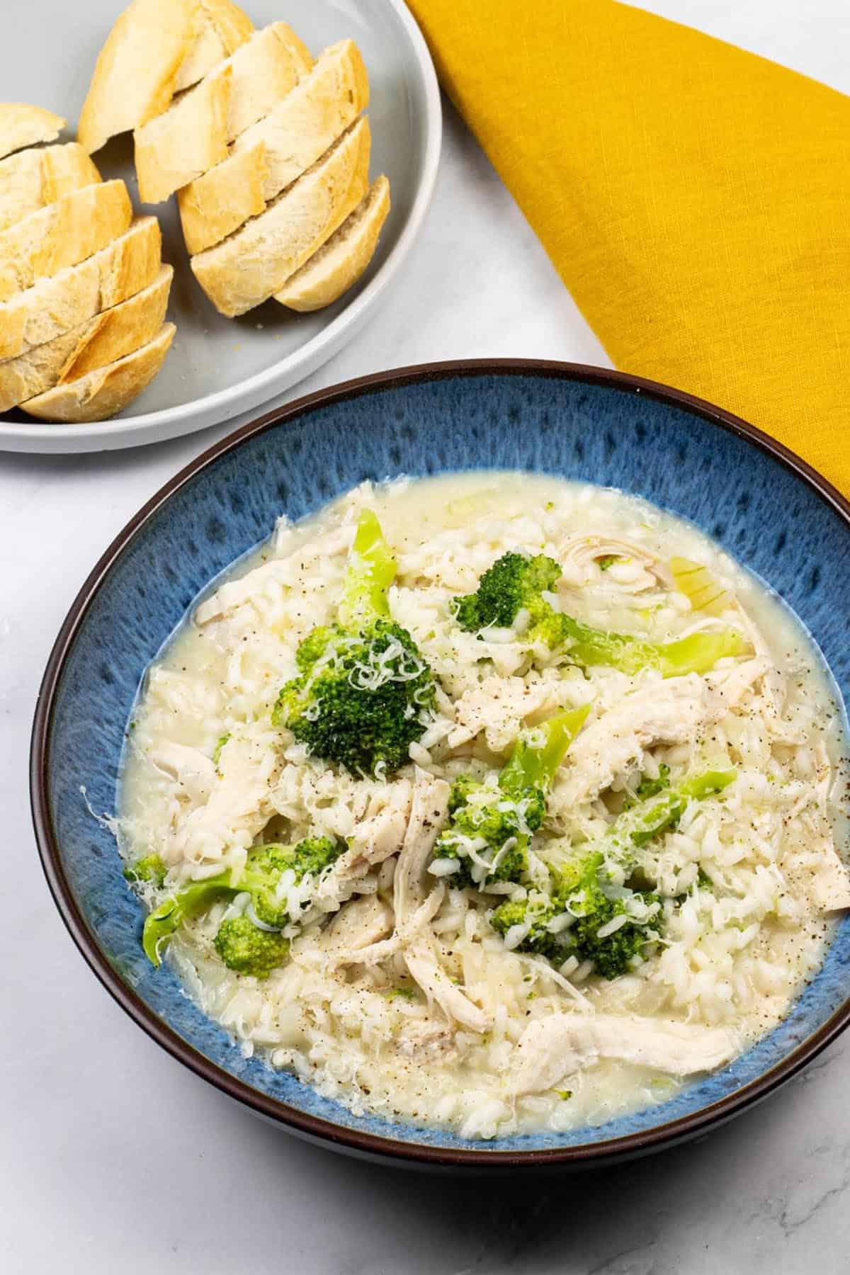Chicken and broccoli risotto in a blue patterned bowl with a plate of sliced baguette and a mustard napkin on the side.