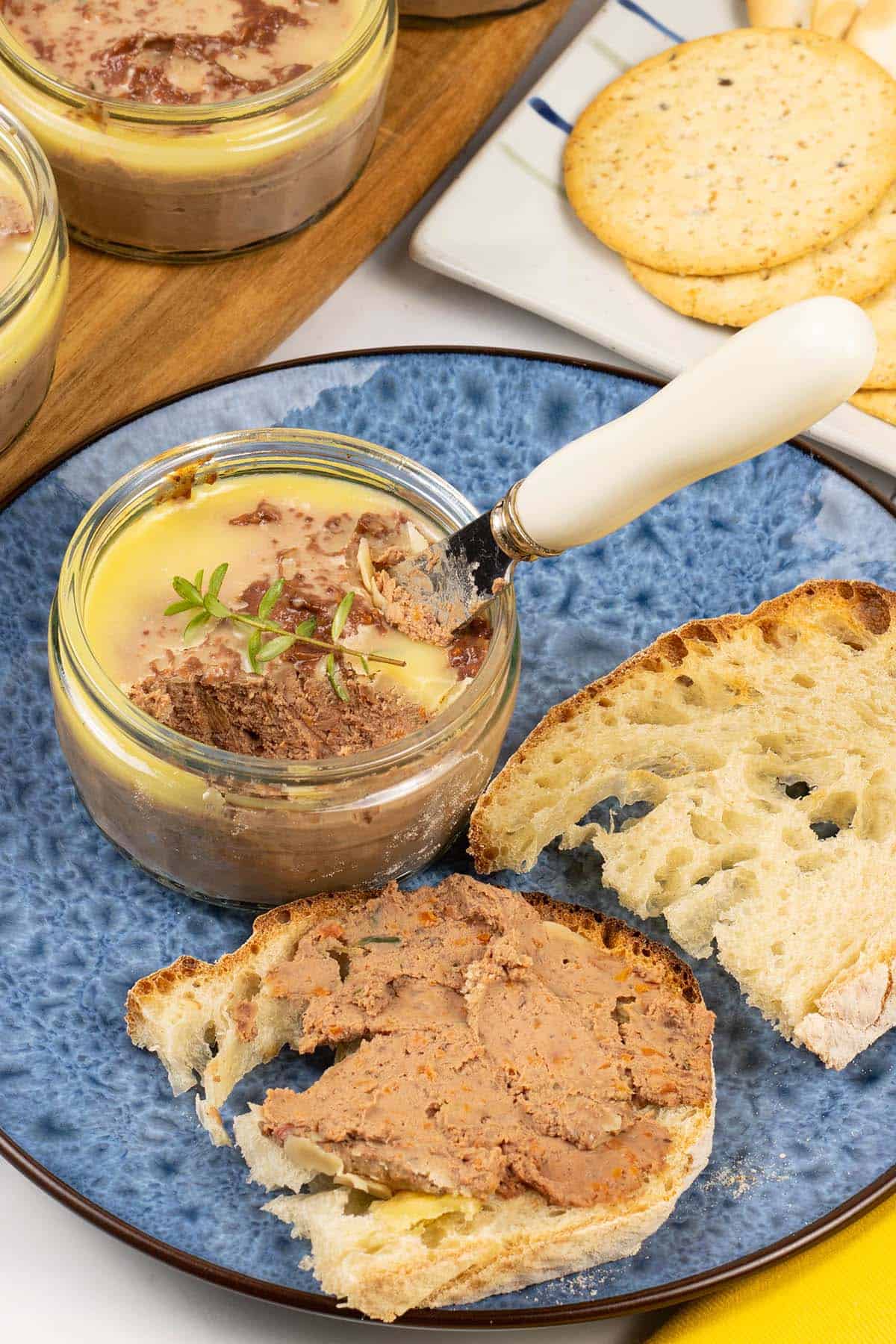 A ramekin of chicken liver pate sitting on a blue patterned side plate with toasted crusty bread. Ramekin dishes of pate and a plate of crackers in the background.