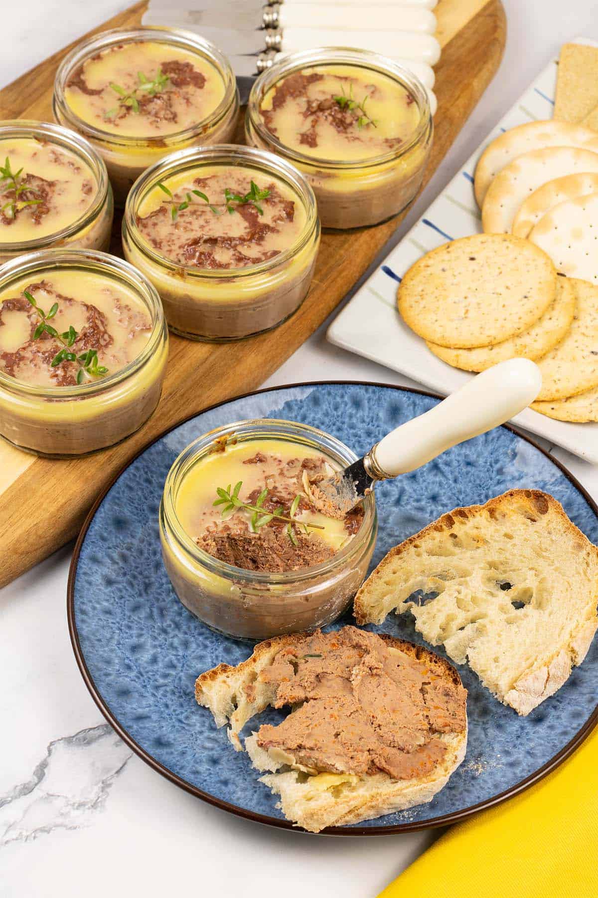 A ramekin of chicken liver pate sitting on a blue patterned side plate with toasted crusty bread. Ramekin dishes of pate and a plate of crackers in the background.