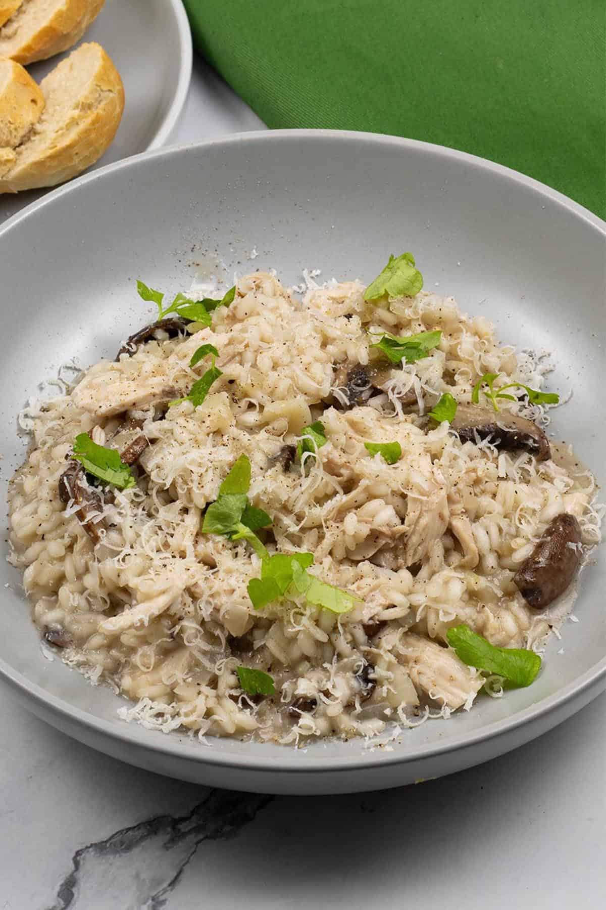 Chicken and mushroom risotto in a grey bowl with a plate of sliced crusty baguette and a green napkin on the side.