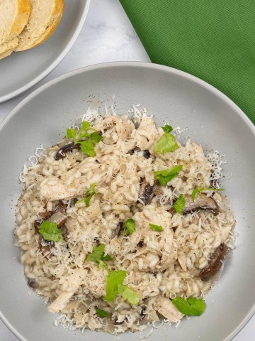 Chicken and mushroom risotto in a grey bowl with a plate of sliced crusty baguette and a green napkin on the side.