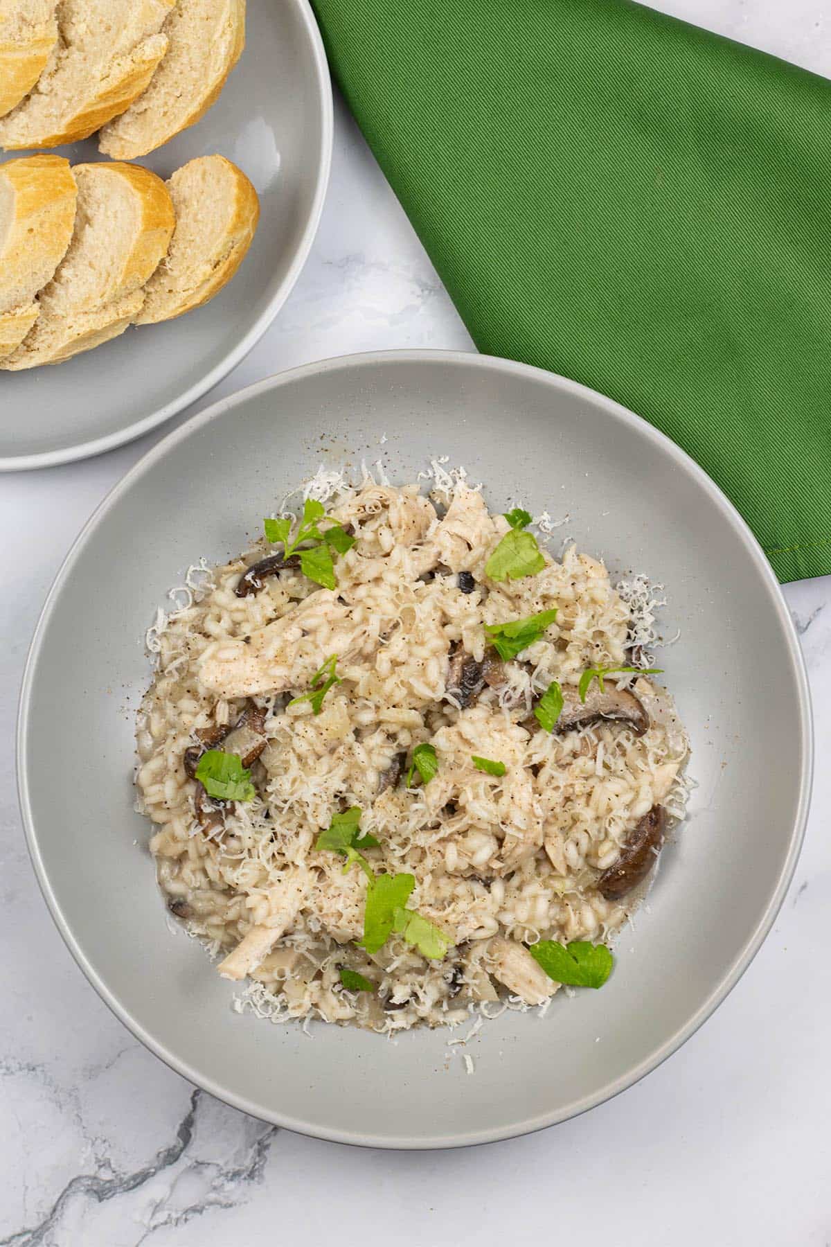 Chicken and mushroom risotto in a grey bowl with a plate of sliced crusty baguette and a green napkin on the side.