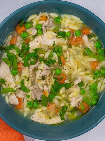 A portion of chicken orzo soup in a teal bowl with an orange napkin on the side.