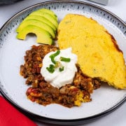 A portion of chilli cornbread pie on a black and white patterned dinner plate, garnished with some sour cream, chopped jalapenos and sliced avocado on the side.