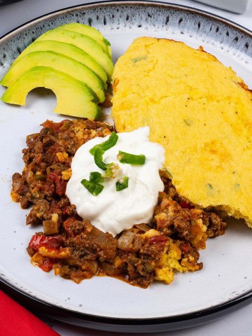 A portion of chilli cornbread pie on a black and white patterned dinner plate, garnished with some sour cream, chopped jalapenos and sliced avocado on the side.