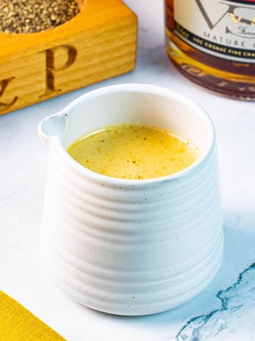 Creamy peppercorn sauce in a small ribbed white jug with a pot of crushed peppercorns and bottle of brandy in the background.