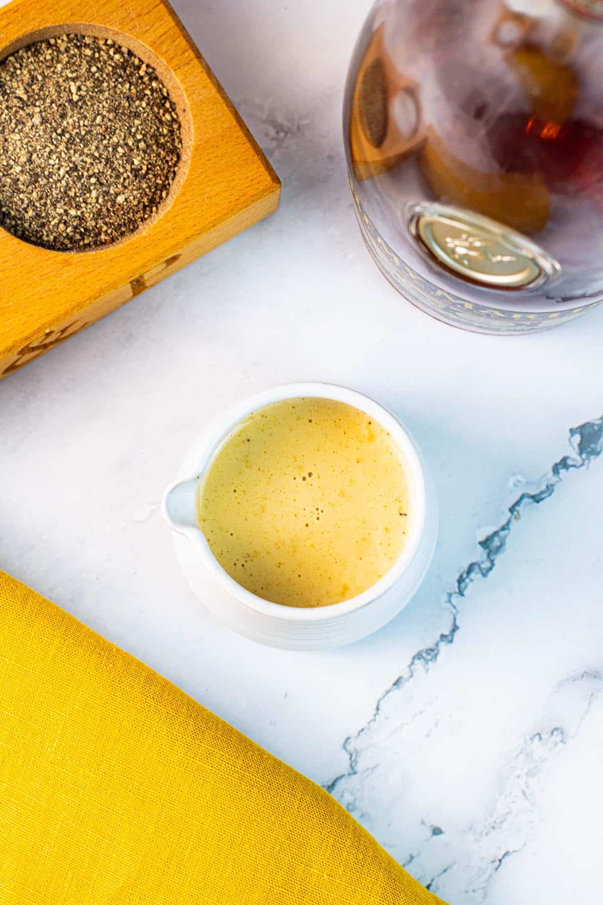 Creamy peppercorn sauce in a small ribbed white jug with a pot of crushed peppercorns and bottle of brandy in the background.