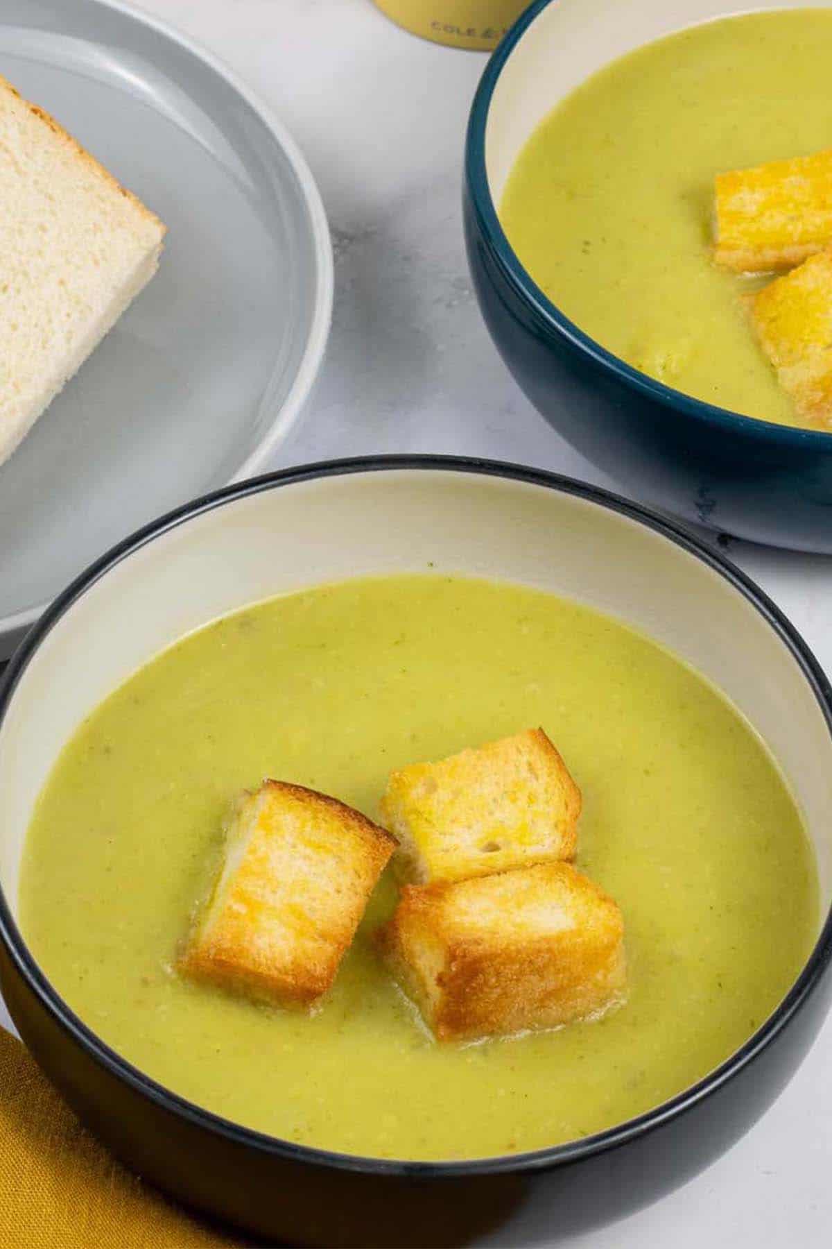 Two bowls of leek and potato soup garnished with croutons and a plate of sliced bread on the side.