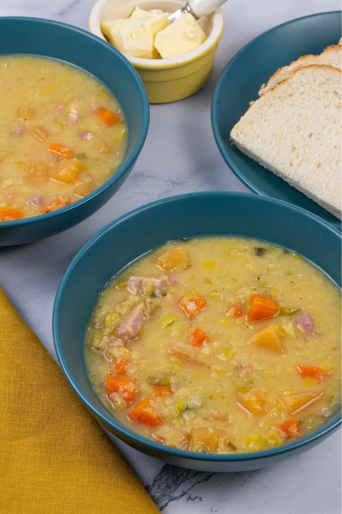 Two portions of lentil soup in teal bowls with sliced bread and a small yellow dish of butter on the side.
