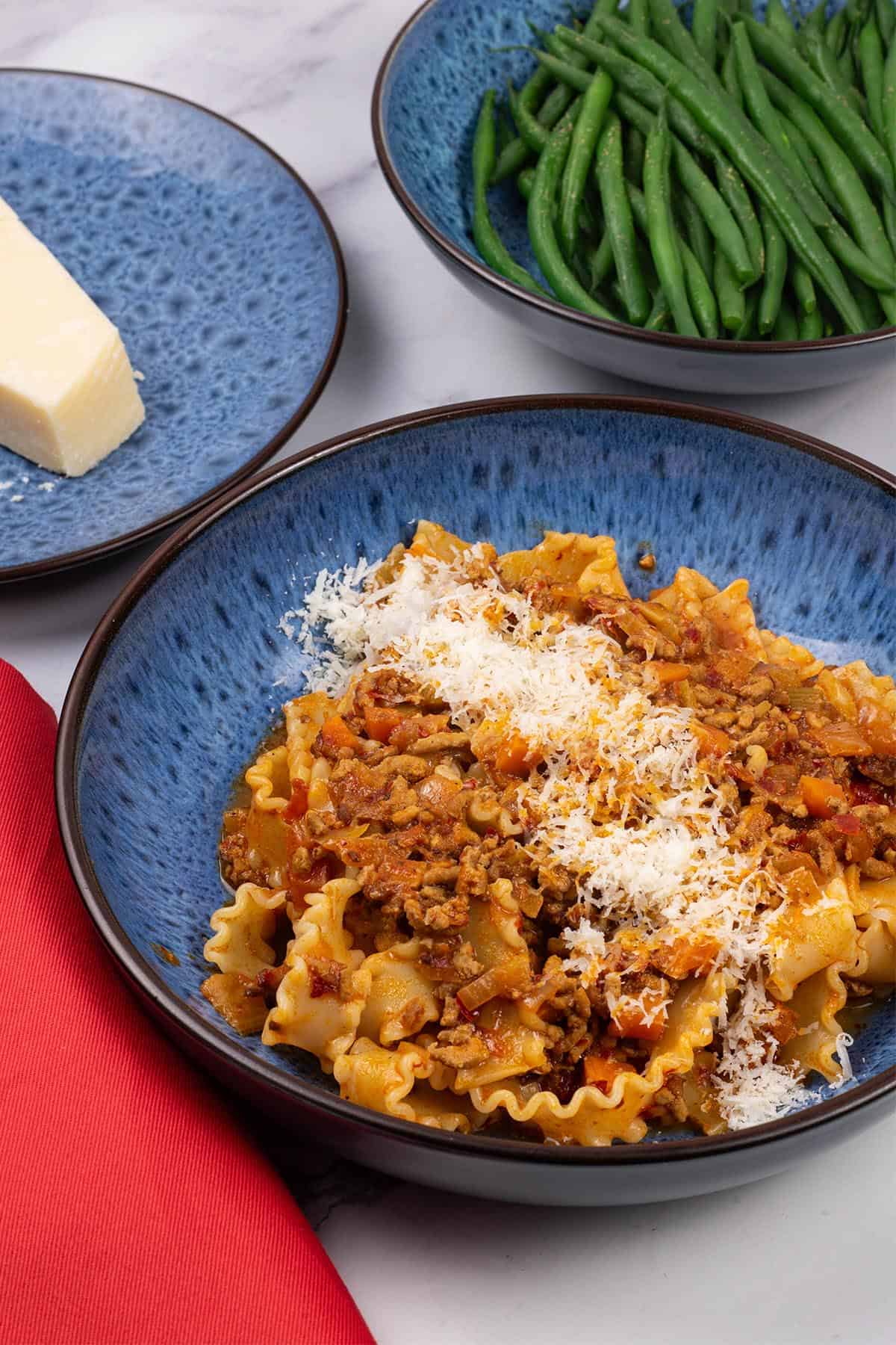 A portion of 'nduja pasta mafalde in a blue patterned bowl with a bowl of green beans and plate with parmesan on the side.