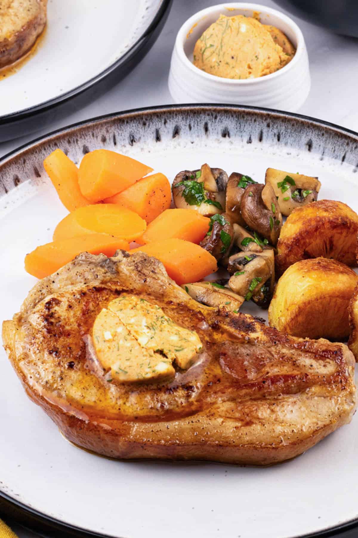 A pork chop with tomato and smoked paprika butter on a black and white dinner plate with carrots, garlic mushrooms and roast potatoes.