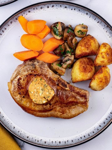 A pork chop with tomato and smoked paprika butter on a black and white dinner plate with carrots, garlic mushrooms and roast potatoes.