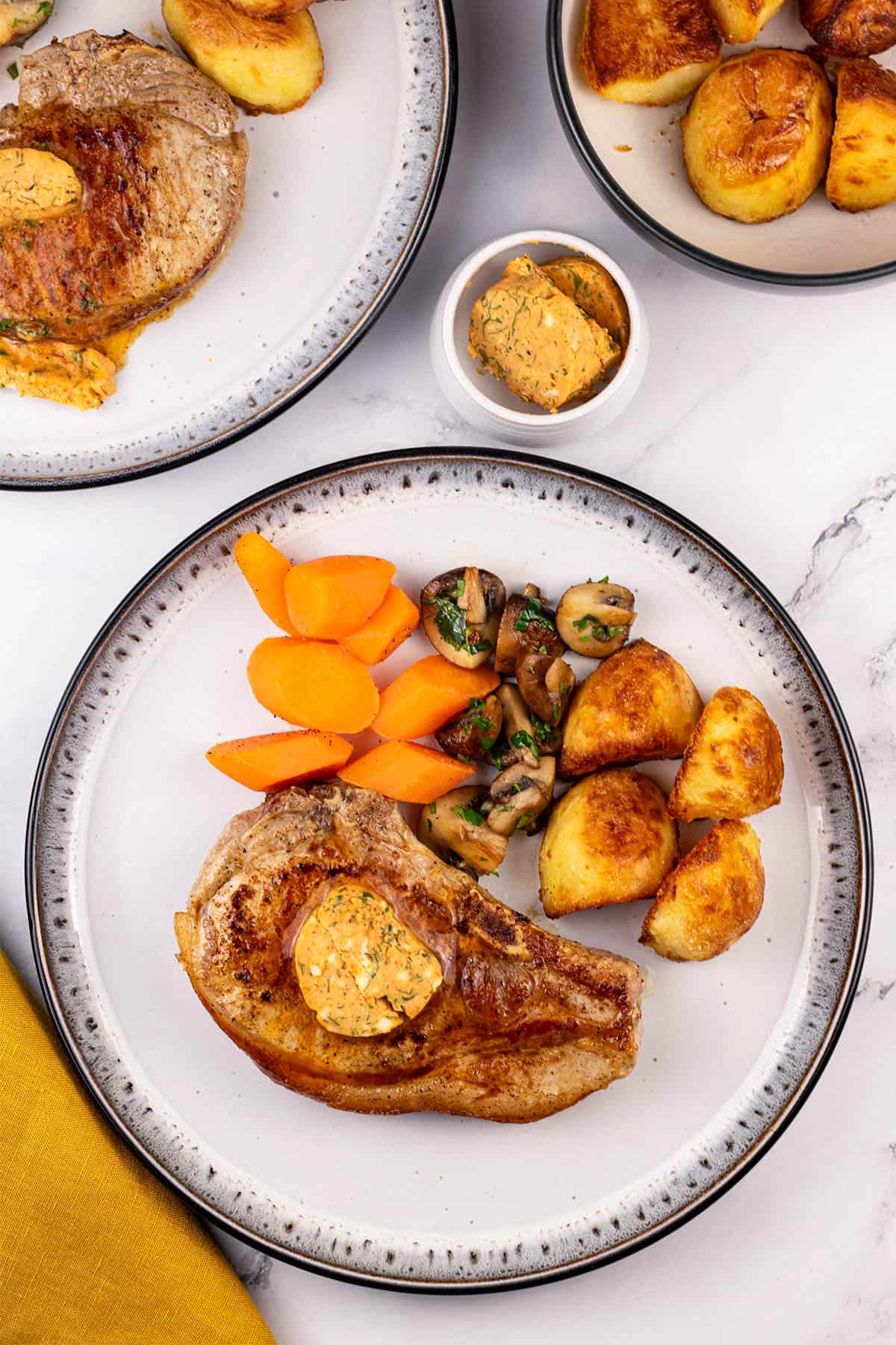 Two portions of pork chops with tomato and smoked paprika butter on dinner plates with carrots, garlic mushrooms and roast potatoes.