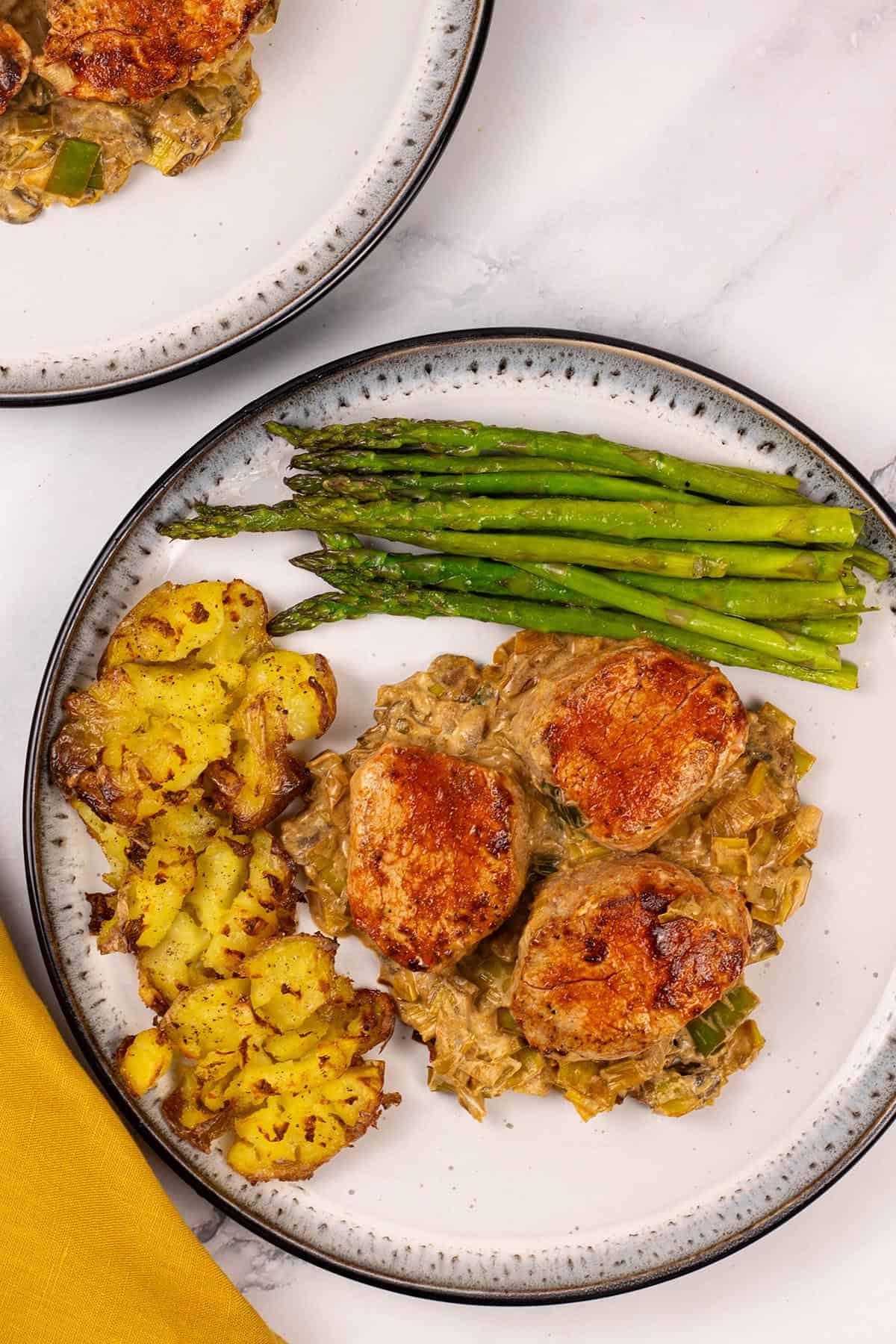 Two portions of pork fillet with mushroom and leek sauce on black and white patterned dinner plates with roast smashed baby potatoes and sauteed asaparagus on the side.