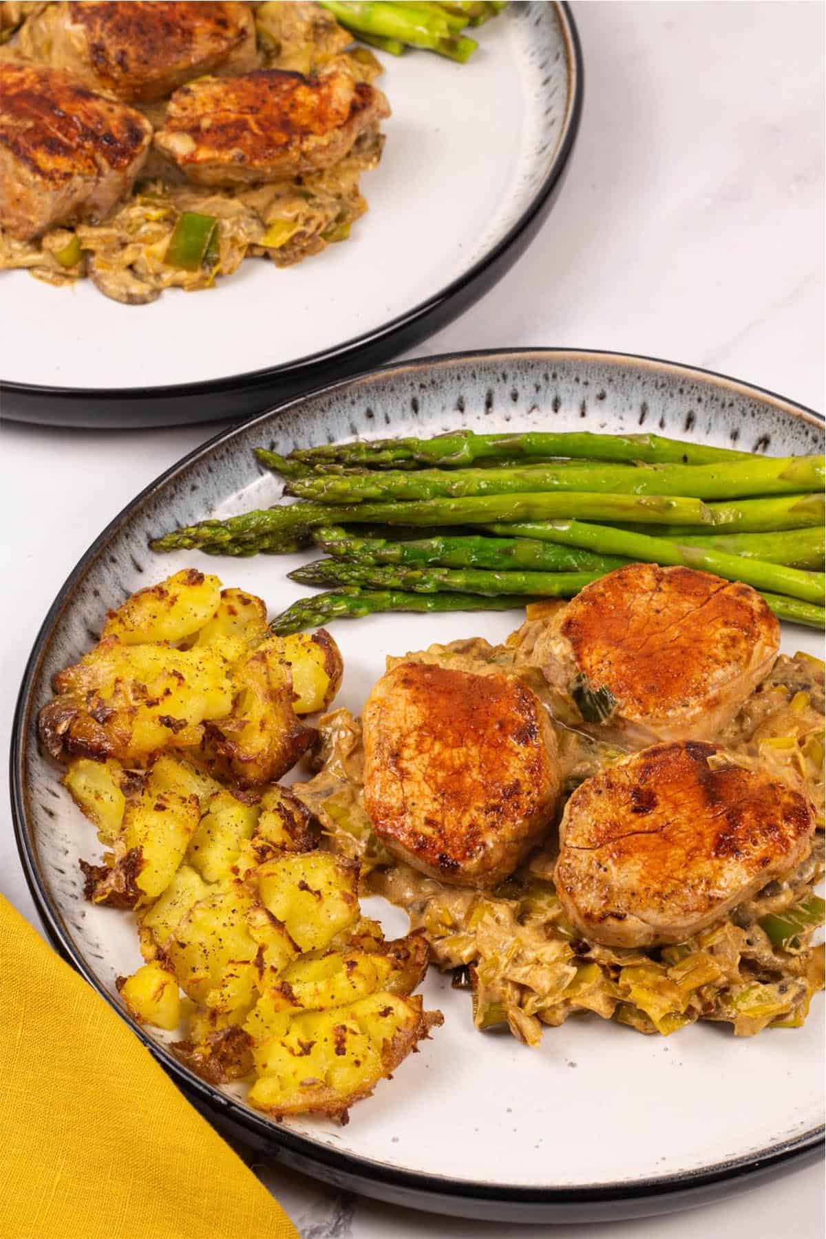Two portions of pork fillet with mushroom and leek sauce on black and white patterned dinner plates with roast smashed baby potatoes and sauteed asaparagus on the side.