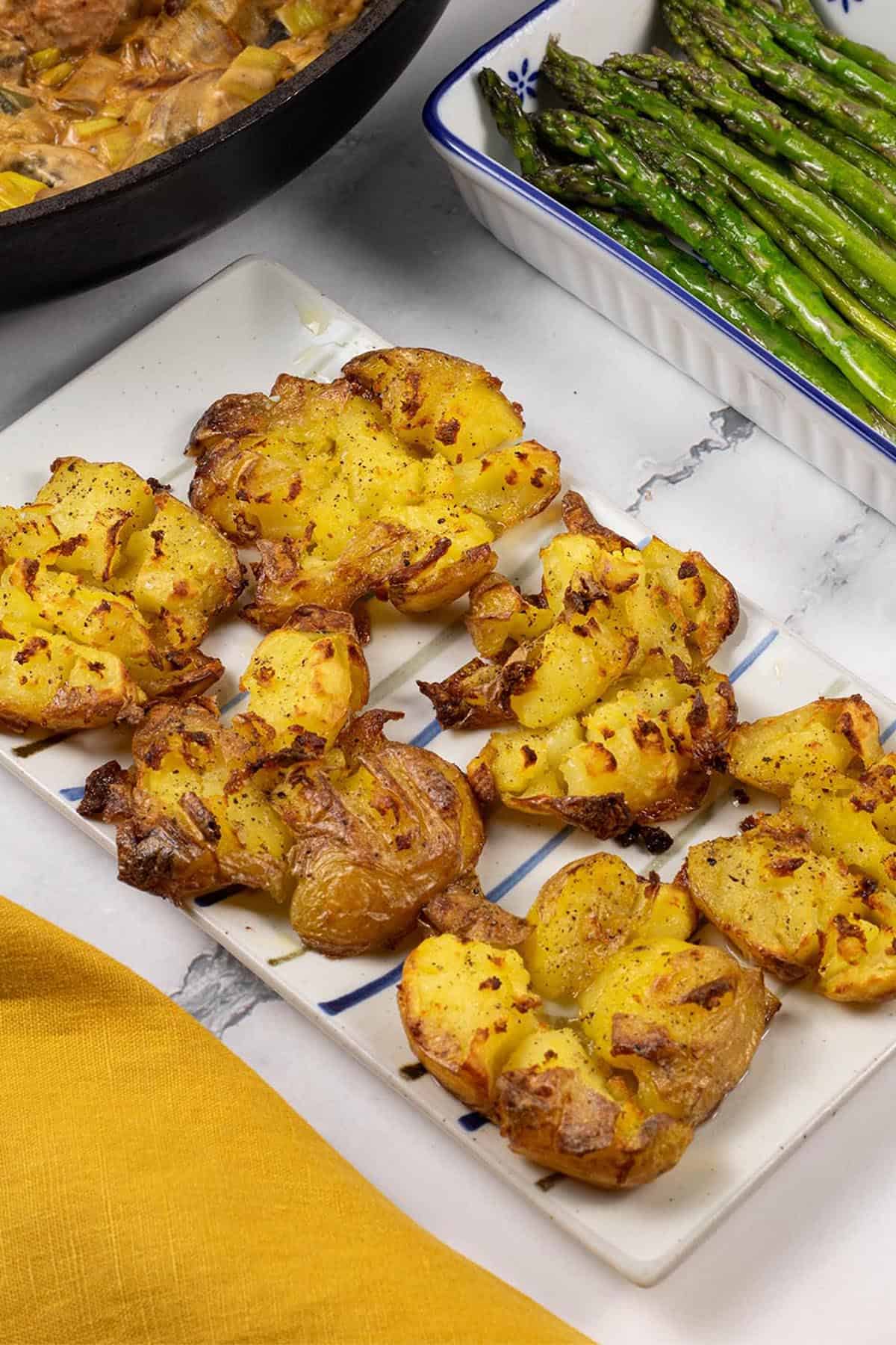 Roast smashed baby potatoes on a white striped platter and pork fillet with mushroom and leek sauce, and sautéed asparagus in the background.