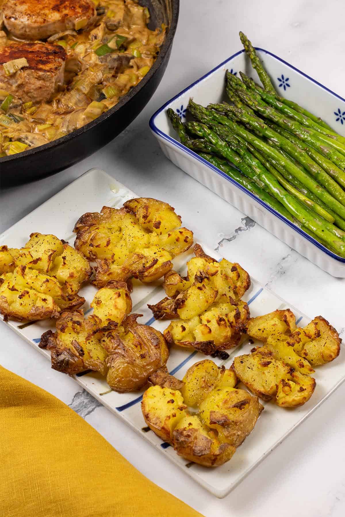 Roast smashed baby potatoes on a white striped platter and pork fillet with mushroom and leek sauce, and sautéed asparagus in the background.