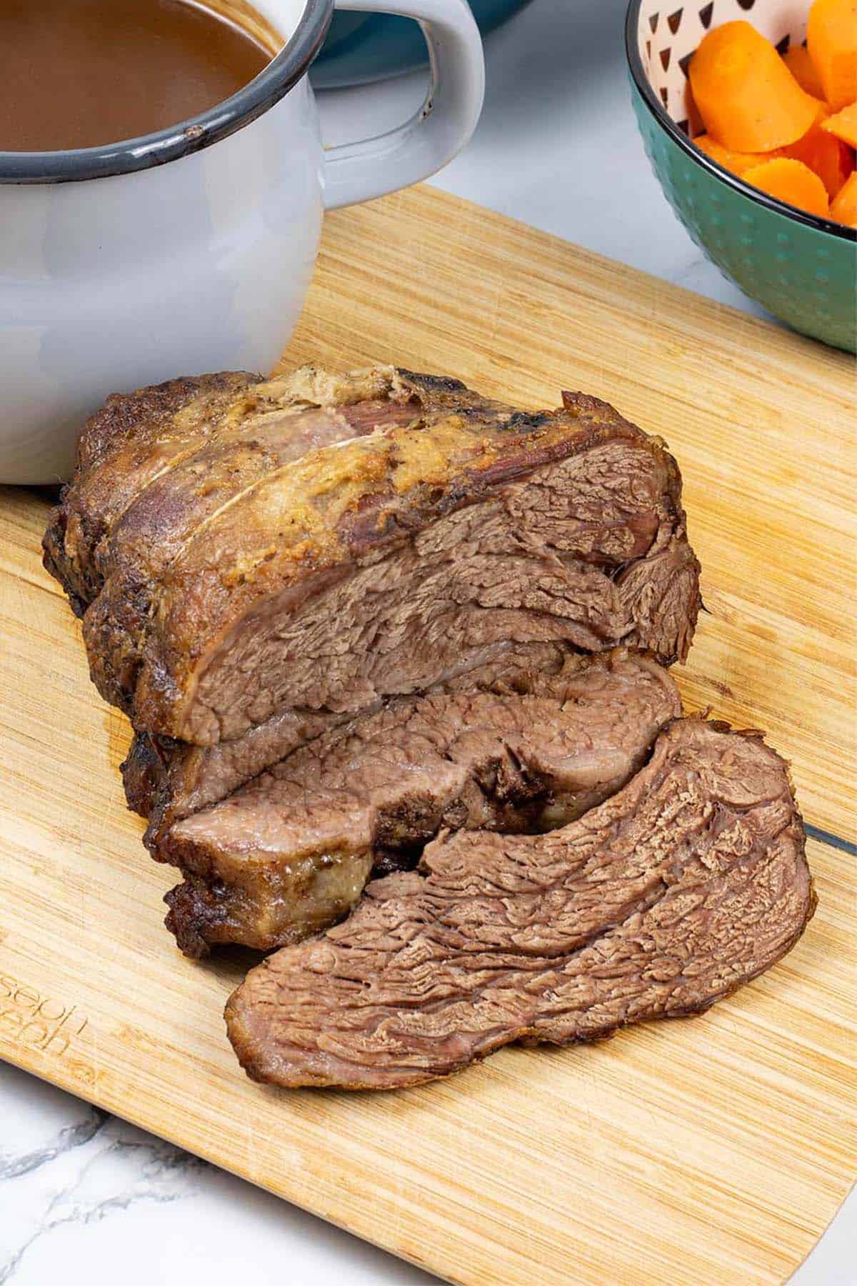 Slow cooker beef pot roast sitting on wooden chopping board with jug of gravy and a bowl of carrots on the side.