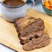 Slow cooker beef pot roast sitting on wooden chopping board with jug of gravy and a bowl of carrots on the side.