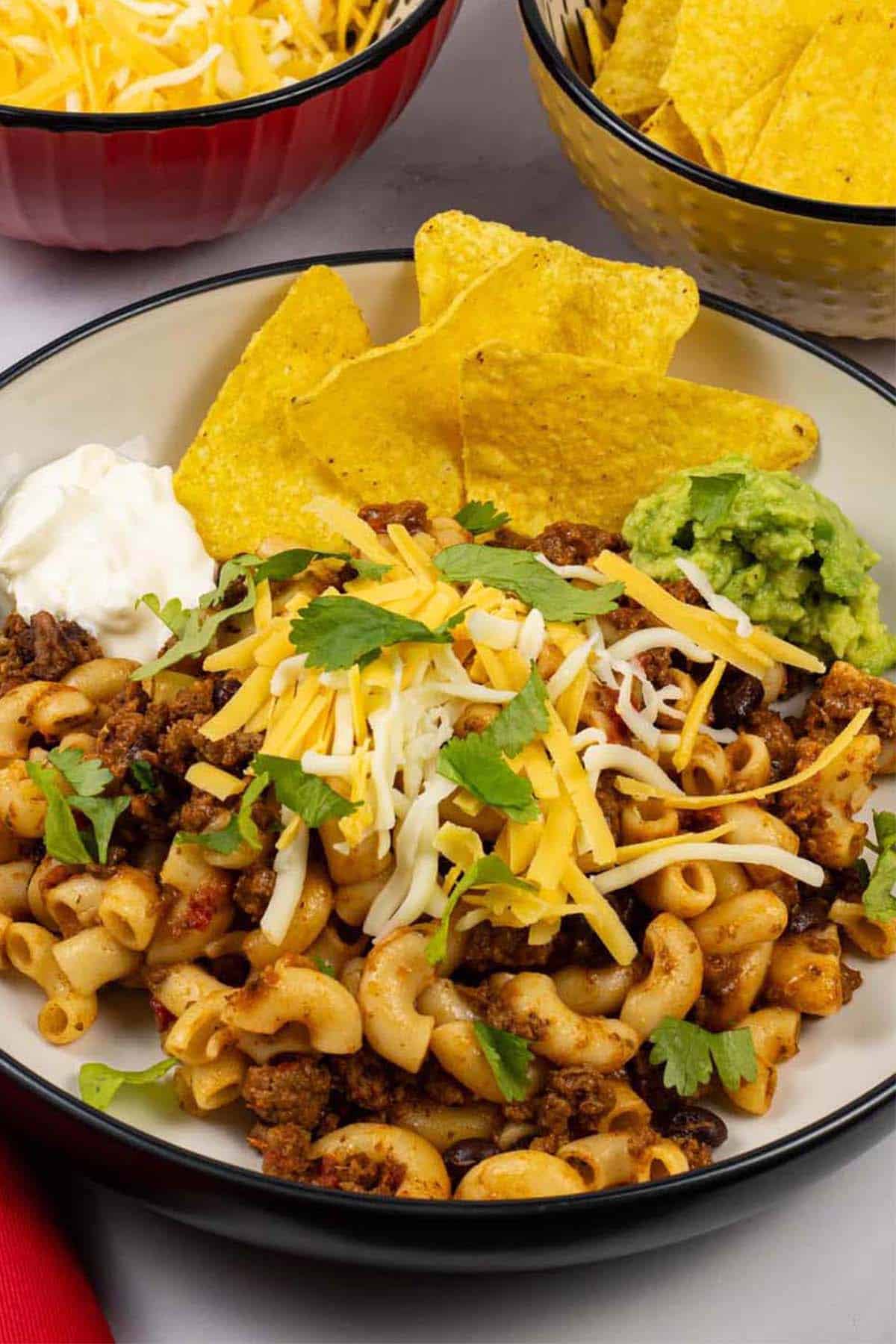 A portion of slow cooker cheesy chilli mac in a black and cream bowl with bowls of grated cheese and tortilla crisps on the side.