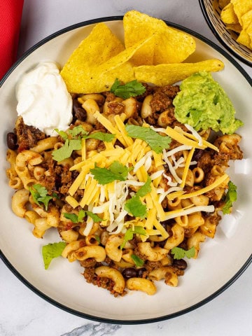 A portion of slow cooker cheesy chilli mac in a black and cream bowl garnished with grated cheese, tortilla crisps, sour cream and guacamole.