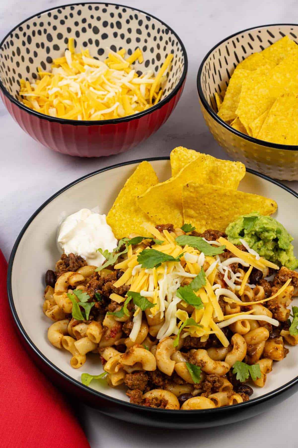 A portion of slow cooker cheesy chilli mac in a black and cream bowl with bowls of grated cheese and tortilla crisps on the side.
