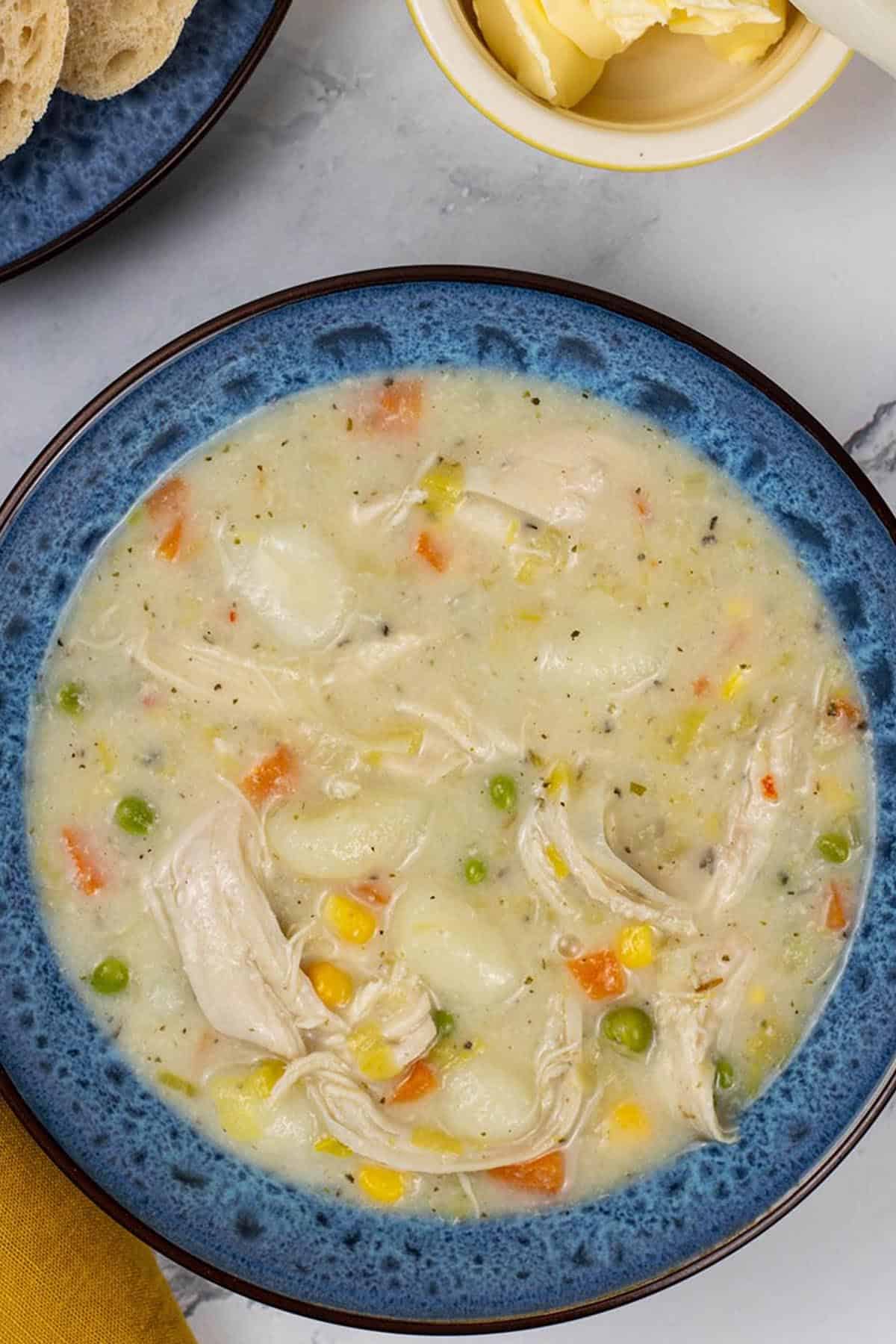 A portion of slow cooker chicken and vegetable gnocchi soup in a blue patterned bowl with a plate of sliced crusty bread and small dish of butter on the side.