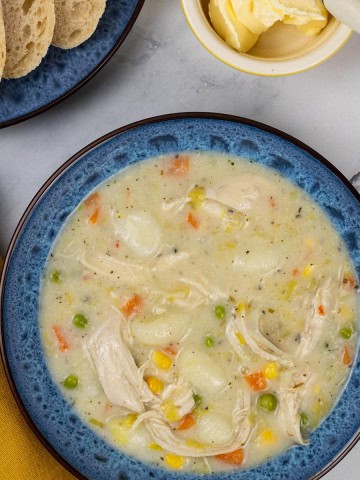 A portion of slow cooker chicken and vegetable gnocchi soup in a blue patterned bowl with a plate of sliced crusty bread and small dish of butter on the side.