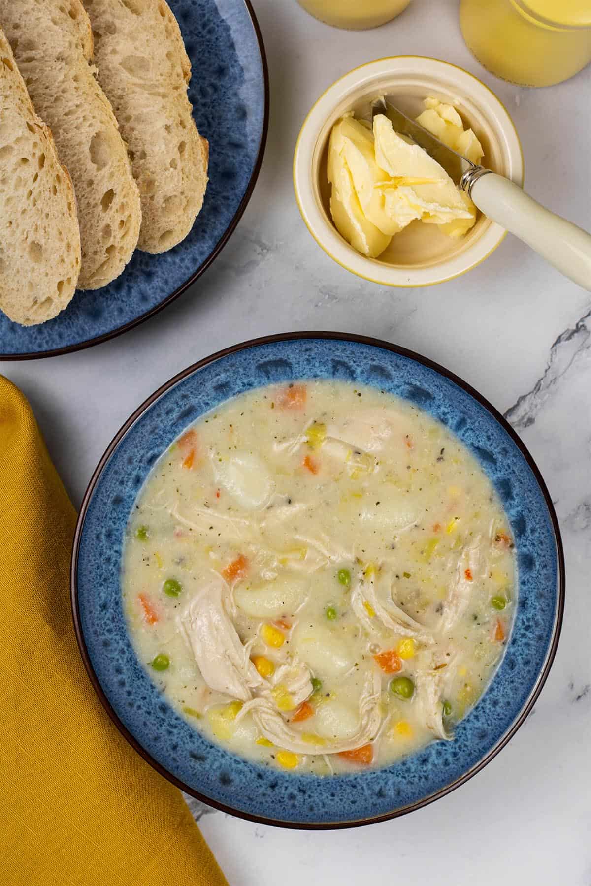 A portion of slow cooker chicken and vegetable gnocchi soup in a blue patterned bowl with a plate of sliced crusty bread and small dish of butter on the side.