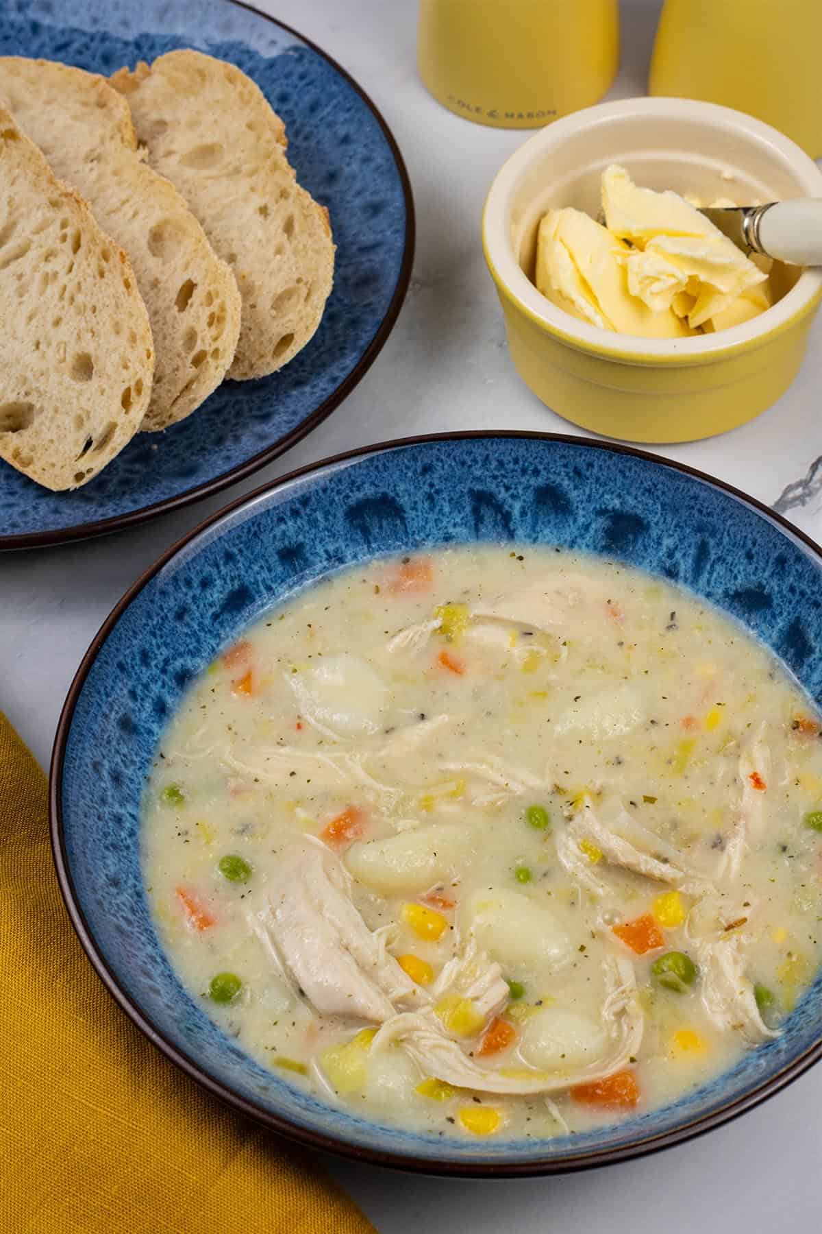 A portion of slow cooker chicken and vegetable gnocchi soup in a blue patterned bowl with a plate of sliced crusty bread and small dish of butter on the side.
