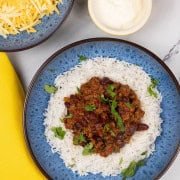 A portion of slow cooker chilli con carne on a bed of rice in a blue patterned bowl, with bowls of grated cheese and sour cream on the side.
