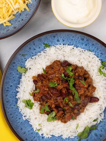 A portion of slow cooker chilli con carne on a bed of rice in a blue patterned bowl, with bowls of grated cheese and sour cream on the side.