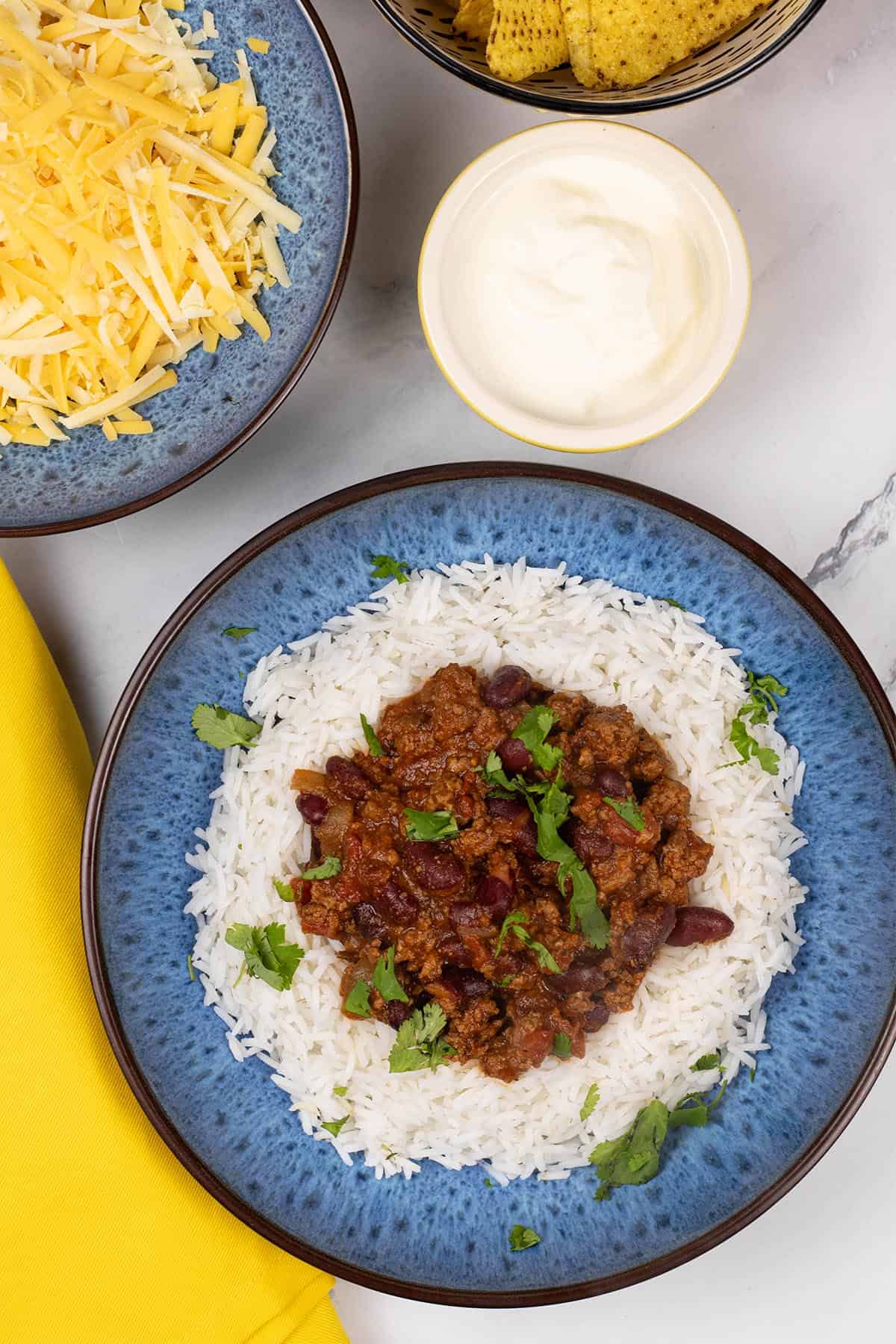 A portion of slow cooker chilli con carne on a bed of rice in a blue patterned bowl, with bowls of grated cheese, sour cream and tortilla chips on the side.