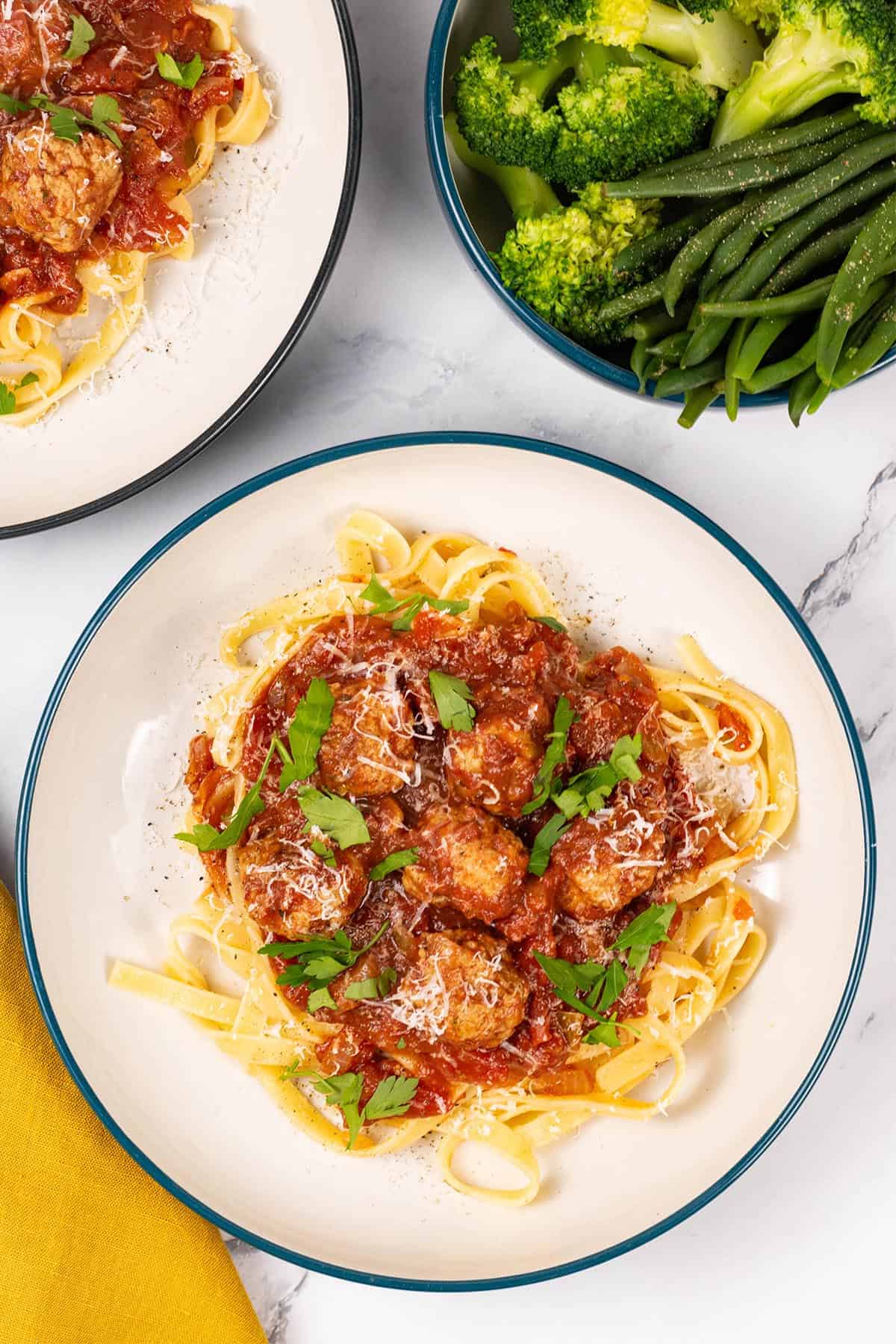Two portions of spicy sausage meatball pasta in pasta bowls with a bowl of quick cook broccoli and quick cook green beans on the side.