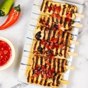 Sweet chilli halloumi skewers on a striped platter with a small dish of sauce on the side.
