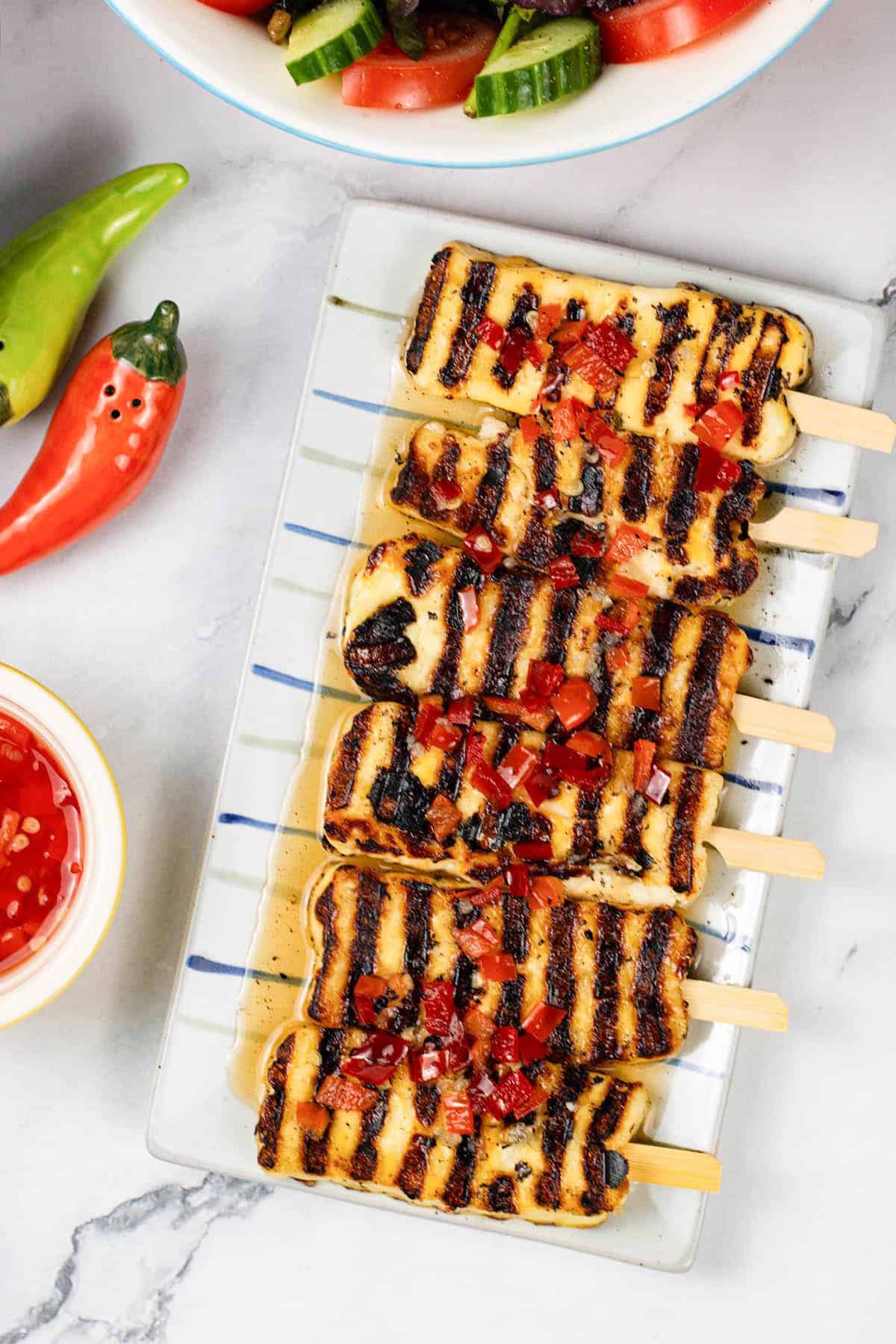 Sweet chilli halloumi skewers on a striped platter with a small dish of sauce and a bowl of salad on the side.