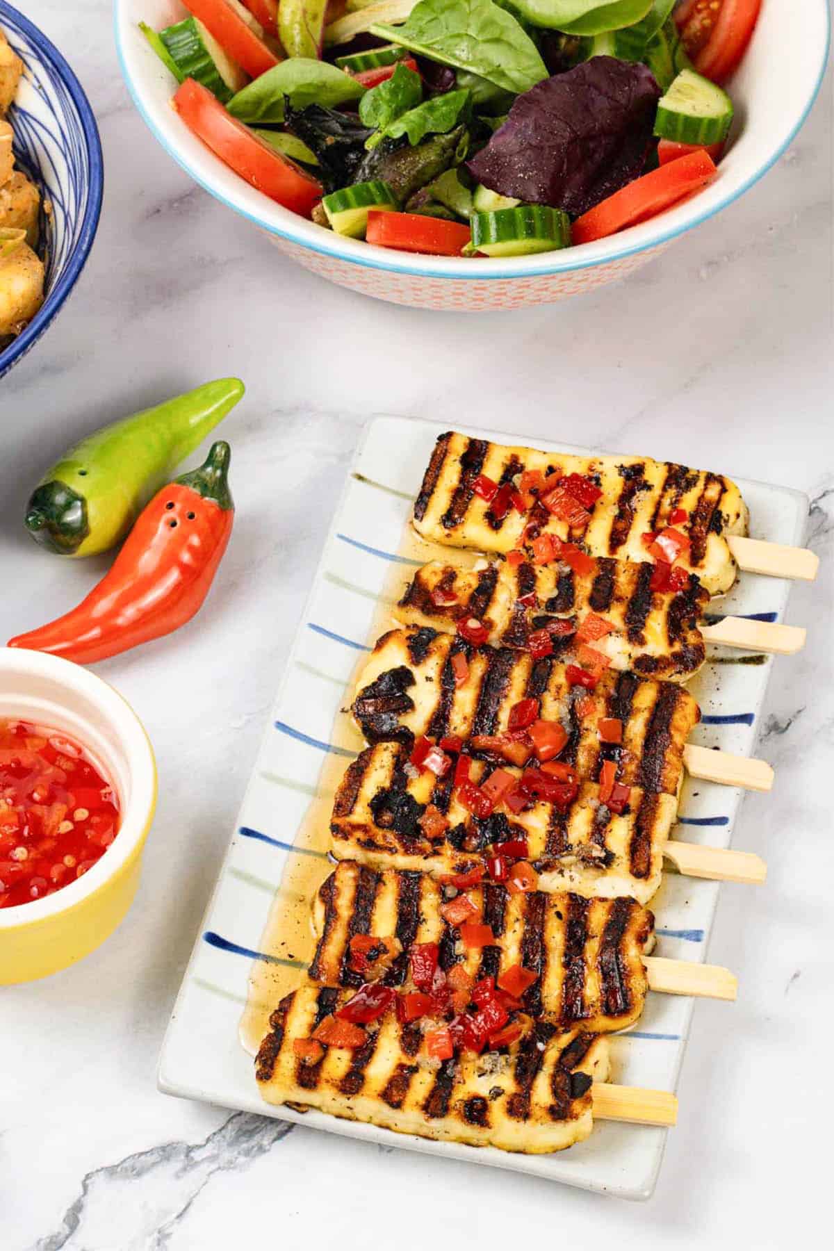 Sweet chilli halloumi skewers on a striped platter with a small dish of sauce and a bowl of salad on the side.