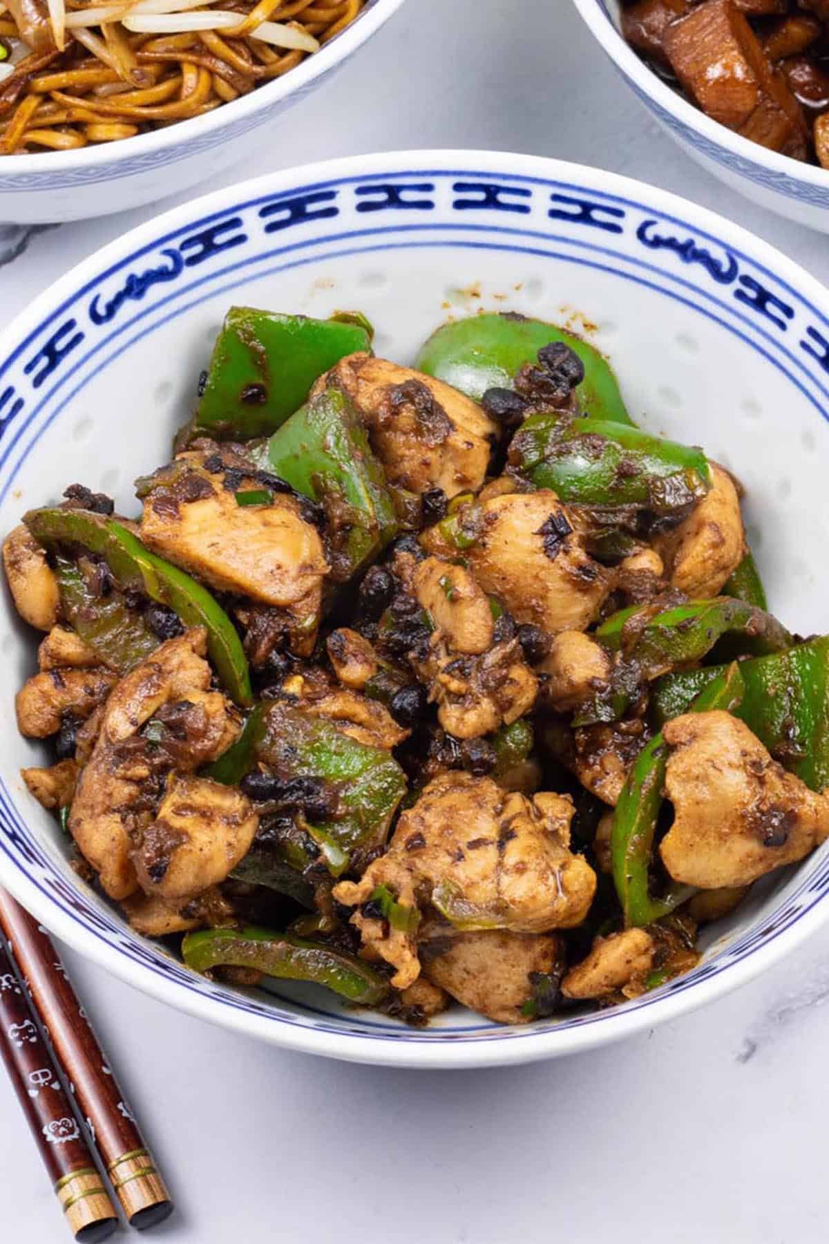 Chicken with green peppers and black bean sauce in a blue and white oriental bowl, with bowls of fried noodles and Shanghai pork belly and chopsticks on the side.