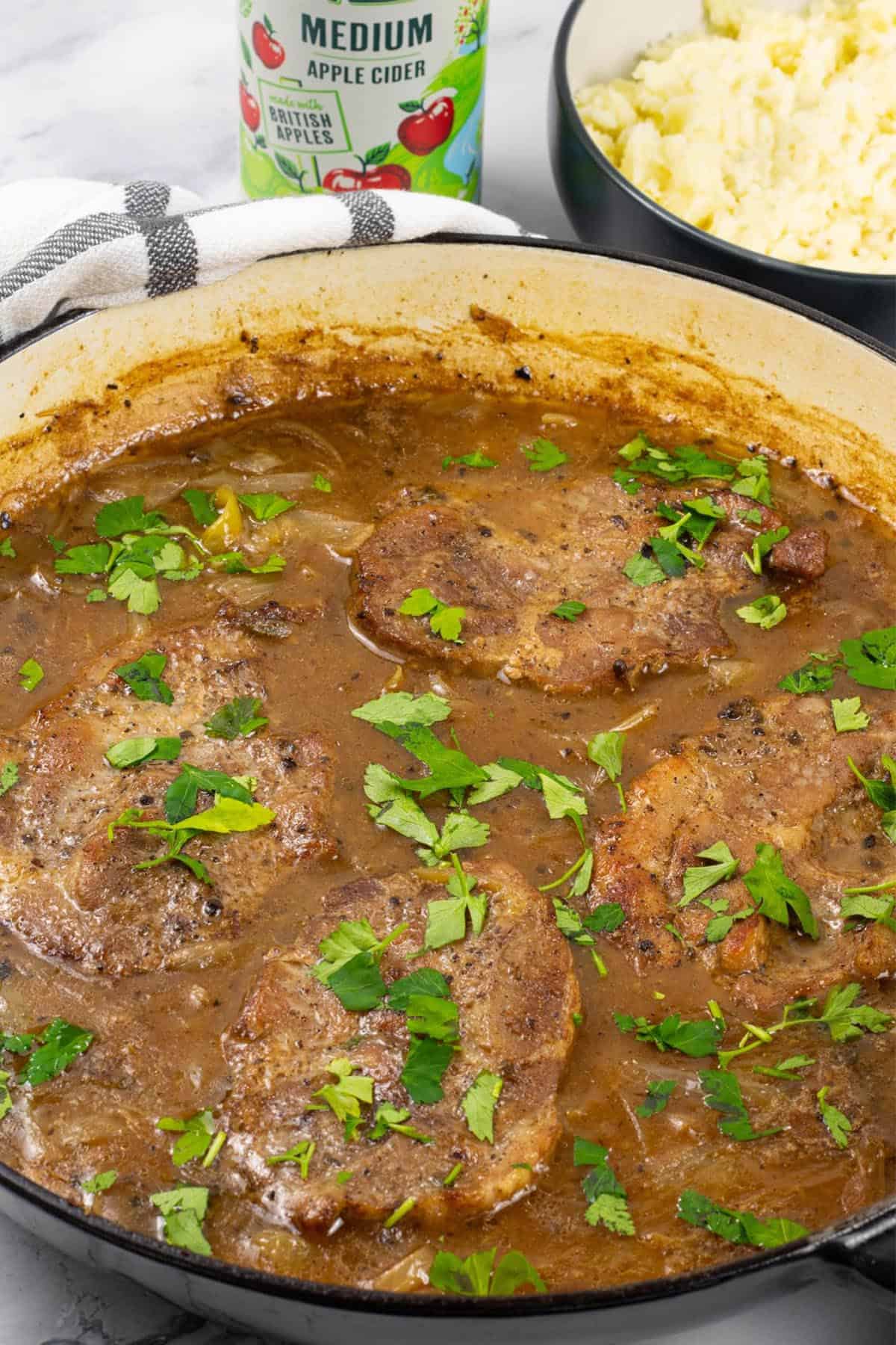 Cider braised pork steaks with onions in a large shallow round casserole, with a bowl of mashed potatoes and can of cider on the side.